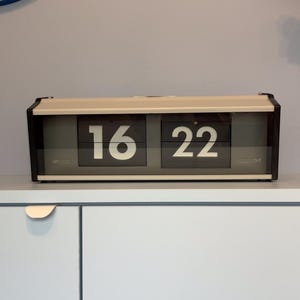 Può includere: Un orologio a scatto in stile retrò che mostra l'ora 16:22. L'orologio ha una cornice nera, una parte superiore color crema e numeri bianchi su pannelli neri. È posto su un mobile blu chiaro.