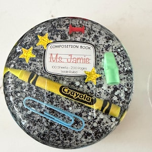 May include: A round, speckled resin coaster featuring school-themed elements. Includes a yellow Crayola crayon, a blue paperclip, a green eraser, and a composition book label that reads "Ms. Jamie". Gold star accents complete the design.