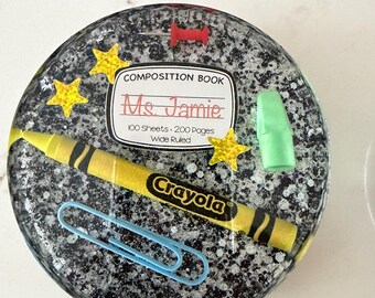 May include: A round, speckled resin coaster featuring school-themed elements. Includes a yellow Crayola crayon, a blue paperclip, a green eraser, and a composition book label that reads "Ms. Jamie". Gold star accents complete the design.