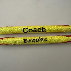 May include: Two yellow glitter pens with black text. One pen reads "Coach" and the other reads "Brooke". The pens have black tips and silver metal accents. Red accents are visible on the pens.