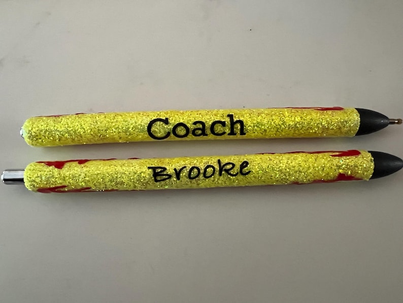 May include: Two yellow glitter pens with black text. One pen reads "Coach" and the other reads "Brooke". The pens have black tips and silver metal accents. Red accents are visible on the pens.
