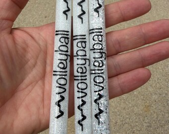 May include: Three white glitter pens with black ink accents. Each pen features a black graphic of a volleyball, a heartbeat line, and the word volleyball printed vertically in black text. The pens have silver click-top mechanisms and black tips.