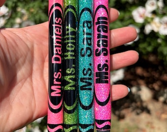 May include: Four retractable pens designed to resemble crayons, featuring glittery barrels in pink, green, teal, and bright pink. Each pen has black accents and is personalized with black text reading Mrs. Daniels, Ms. Holly, Ms. Sara, and Ms. Sarah.