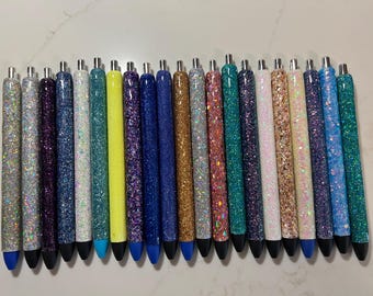 May include: A collection of glitter pens in various colors, including silver, purple, blue, yellow, gold, pink, and teal. Each pen has a black tip and a silver metal top. The pens are arranged in a row.