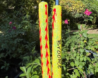 May include: Two yellow glitter pens with red stitching detail, resembling baseballs. The word "Brooke" is written vertically on one pen. The pens are held in front of a green, leafy background.