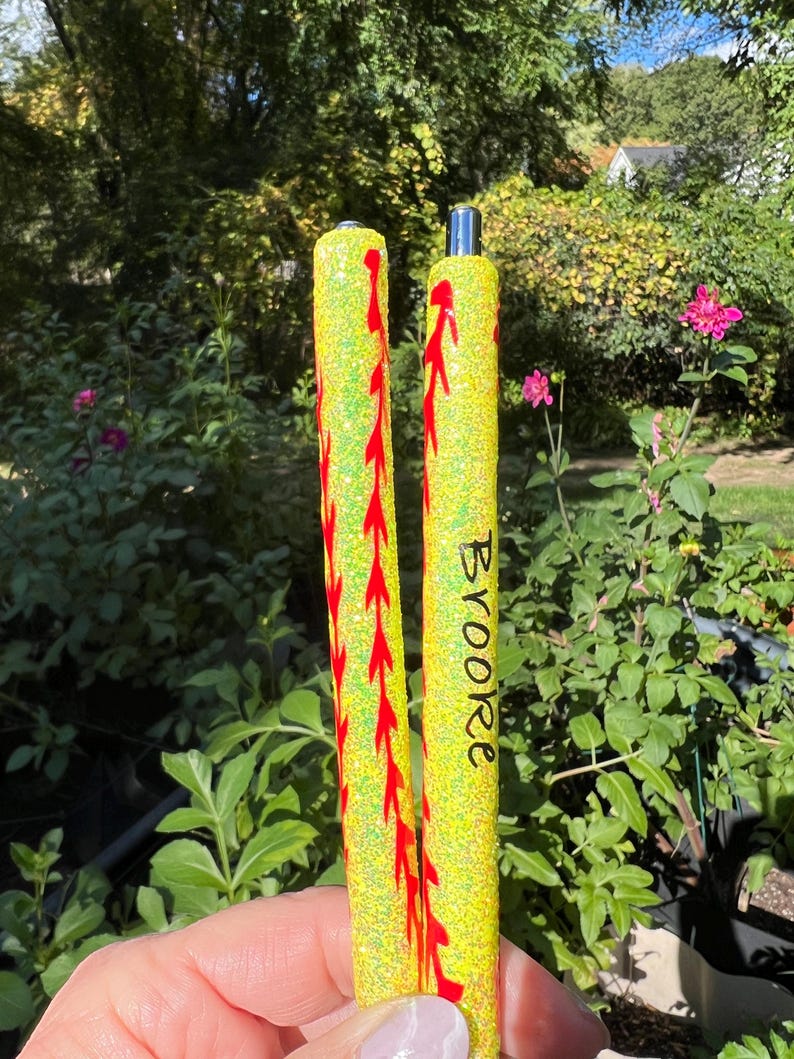 May include: Two yellow glitter pens with red stitching detail, resembling baseballs. The word "Brooke" is written vertically on one pen. The pens are held in front of a green, leafy background.