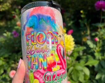 May include: A glitter-covered tumbler with a colorful design. The tumbler features the words "Float Drink Tan Repeat" with a flamingo and cocktail graphic. The background has a watercolor effect with shades of blue, pink, and orange.