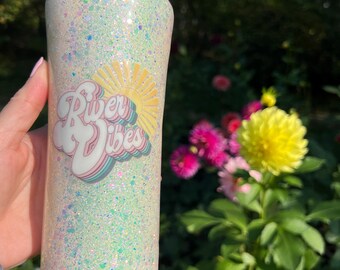 May include: A glitter-covered tumbler with a black lid. The tumbler has a pastel color scheme with a "River Vibes" graphic featuring a sun and clouds. The background includes green foliage and colorful flowers.