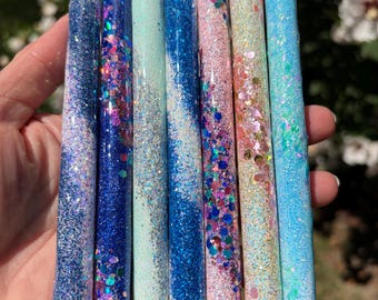 May include: Seven retractable pens with glittery, textured barrels in shades of blue, pink, and white. Each pen features a silver-toned top clicker and a black conical tip. The designs include solid glitter, chunky sequins, and gradient patterns.