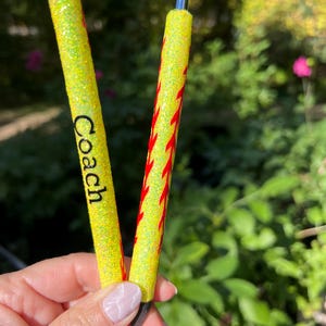 May include: Two yellow glitter pens. One pen has the word "Coach" in black. The other pen has a red flame design. The pens are held in a hand against a blurred green background.