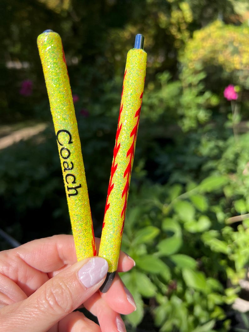 May include: Two yellow glitter pens. One pen has the word "Coach" in black. The other pen has a red flame design. The pens are held in a hand against a blurred green background.