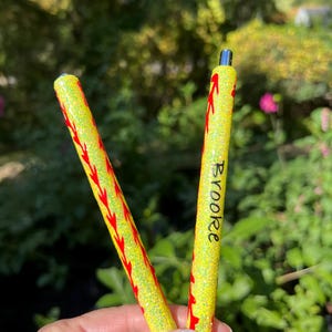 May include: Two yellow glitter pens with red stitching-like designs. One pen has the name "Brooke" written in black. The pens are held in front of a blurred green and pink background.