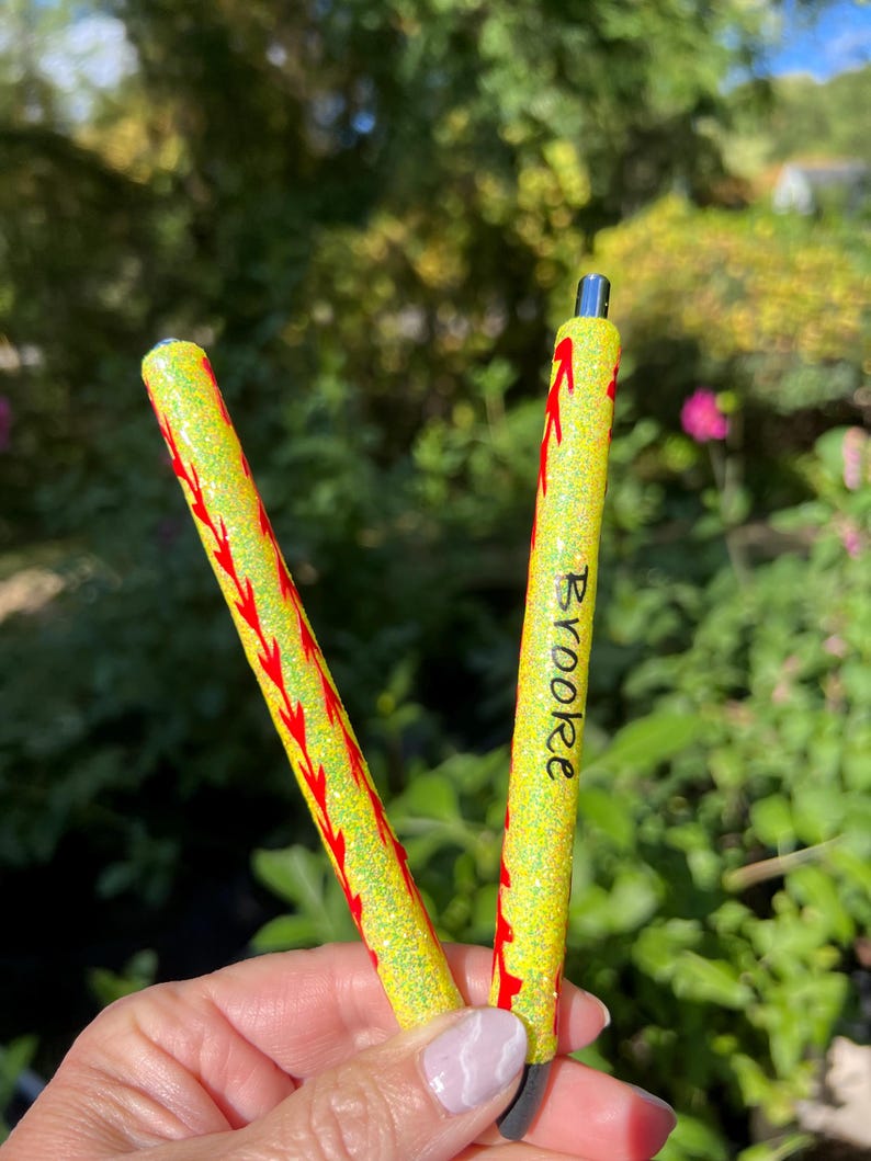 May include: Two yellow glitter pens with red stitching-like designs. One pen has the name "Brooke" written in black. The pens are held in front of a blurred green and pink background.