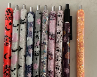 May include: A collection of nine retractable pens featuring various Halloween-themed designs including ghosts, bats, pumpkins, and flowers. The pens have glittery finishes in colors such as orange, white, purple, silver, and pink, with black ink tips.