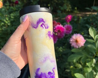 May include: A white travel mug with a black lid, featuring a swirling design of purple and yellow. The tumbler is held in front of a background of green foliage and pink flowers.