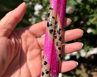 May include: Two glittery pens featuring a vibrant pink and magenta color scheme with gold accents and black leopard print patterns. Each pen has a smooth, shiny finish, a silver-toned top click mechanism, and a black conical tip at the base.