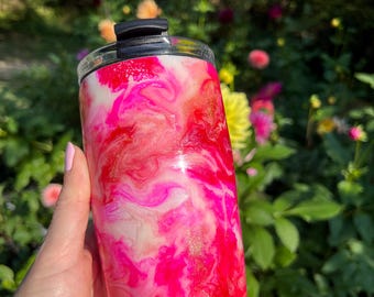 May include: A stainless steel tumbler with a vibrant, swirled design in shades of pink, white, and gold. The tumbler has a black lid and a clear rim. The background is a blurred view of green foliage and colorful flowers.