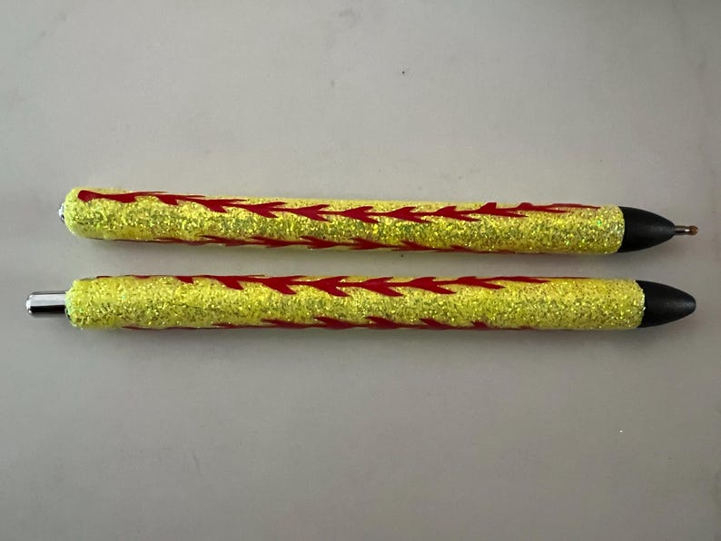 May include: Two glitter pens with yellow barrels and red arrow designs. The pens have black tips and silver accents. The pens are approximately 6 inches long.