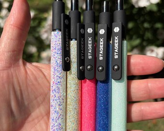 May include: Five retractable pens with black clips and caps, each featuring a distinct glittery barrel in white, gold, pink, blue, and light green. Each black clip is clearly labeled with the brand name STAGEGEEK and the model designation GX3.