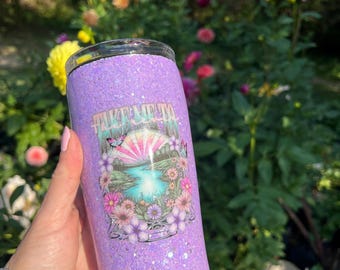 May include: A lavender glitter tumbler with a clear lid. The tumbler features a colorful graphic of a landscape with a waterfall, flowers, and the text "Take Me Away". The tumbler is held in a hand.