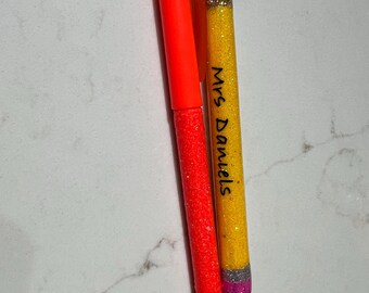 May include: Two pens. One is bright orange with a glittery texture, and the other is yellow with glitter, resembling a pencil. The yellow pen has the text "Mrs. Daniels" written on it. The pencil-shaped pen has a black tip and a silver and pink base.