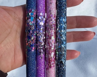 May include: Four glitter pens in shades of blue, purple, and pink. The pens have black tips and silver accents. The glitter is embedded in the pen barrels, creating a sparkling effect. The pens are held in a hand.