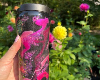 May include: A black travel tumbler with a black lid, featuring a vibrant design of pink and purple swirls with glitter accents. The tumbler is held in a hand, with a blurred garden background.