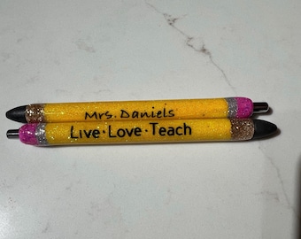 May include: Two yellow glitter pens with black tips and pink and silver accents. One pen reads "Mrs. Daniels" and the other reads "Live Love Teach". The pens are designed to resemble pencils.