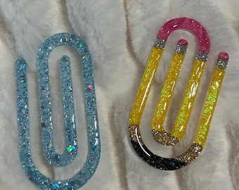 May include: Two resin paper clips. One is light blue with glitter. The other is designed to look like a pencil, with yellow, pink, black, and silver glitter accents. Both are on a white, fluffy surface.