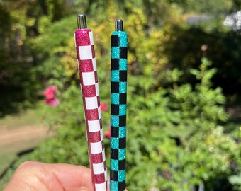 May include: Two cylindrical pens featuring a checkered pattern. One pen displays a magenta glitter and white checkerboard design, while the other pen features a teal glitter and black checkerboard pattern. Both pens have a silver-colored top clicker.