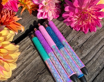 May include: A collection of six colorful pens with glittery, mermaid scale-patterned barrels. The pens are arranged on a weathered wooden surface, surrounded by vibrant pink, orange, and yellow dahlias. The pens have black tips.