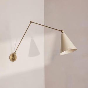 May include: A wall-mounted brass lamp with a cream-coloured, cone-shaped shade. The lamp extends from a circular base and features a long, articulated arm. The shade has small holes near the bottom edge. The lamp is designed to be adjustable.