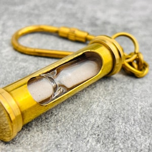May include: A gold-toned metal hourglass keychain featuring a cylindrical frame with a clear glass center containing fine white sand. The hourglass is attached to a sturdy gold-colored metal carabiner clip and a small circular ring for keys.