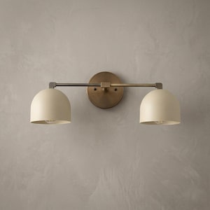 Könnte beinhalten: Eine zweiflammige Wandleuchte mit Vintage-Messing-Finish. Die Leuchte hat zwei cremefarbene, kuppelförmige Schirme und eine kreisförmige Rückplatte. Die Leuchte ist an einer grauen Wand montiert.