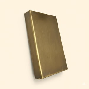 Könnte beinhalten: Ein rechteckiger Gegenstand aus massivem Messing mit gebürsteter Oberfläche. Das Messing hat einen warmen, goldenen Farbton und eine leicht strukturierte Oberfläche. Das Objekt ist abgewinkelt und zeigt seine Dicke und die klaren Linien seiner Kanten.