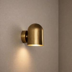 Op de afbeelding: Een messingkleurige wandlamp met een afgeronde, koepelvormige kap. De lamp is aan een muur bevestigd en straalt een warme gloed uit. De lamp heeft een cilindrische basis en een gebogen bovenkant, wat een moderne esthetiek creëert.