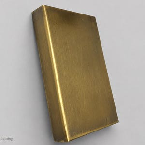 May include: A rectangular brass object with a smooth, metallic gold-toned surface and a visible spine-like edge. The item features a solid, flat construction with subtle surface variations and the text Emberixlighting printed in the bottom corner.