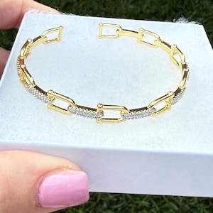 May include: A gold-toned bracelet with a chain link design. The bracelet features rectangular links, with sections of the links encrusted with small, clear gemstones. The bracelet is presented in a white box, suggesting it is a piece of jewelry.
