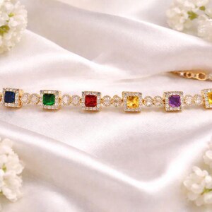 May include: A gold bracelet with seven square-cut gemstones in blue, green, red, yellow, and purple, each surrounded by small clear stones. The bracelet is on a white satin surface, with white flowers in the background.