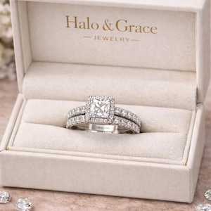 May include: A diamond engagement ring set in a white box. The ring features a square-cut center stone surrounded by a halo of smaller diamonds. The band is also encrusted with diamonds. The box is labeled "Halo & Grace - JEWELRY".