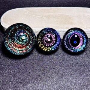 May include: Three glass galaxy-themed plugs in varying sizes. Each plug features a swirling, colorful design with iridescent accents. The plugs are primarily black with shades of blue, purple, red, and yellow. The plugs are displayed on a gray surface.
