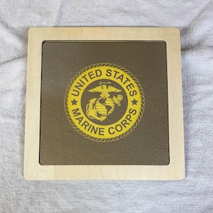 May include: A wooden plaque featuring the United States Marine Corps emblem. The emblem, in yellow, is centered on a brown background and includes an eagle, globe, and anchor design, surrounded by the words "United States Marine Corps". The plaque has a light wood frame.