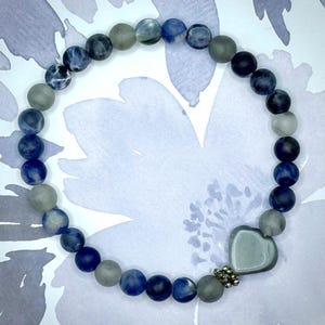 Könnte beinhalten: Ein Armband mit runden blauen, grauen und weißen Perlen. Ein herzförmiger, grauer Anhänger befindet sich in der Mitte des Armbands, akzentuiert durch eine kleine, silberfarbene Perlenansammlung. Das Armband wird vor einem hellblauen Blumengrund gezeigt.