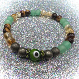 Könnte beinhalten: Ein Perlenarmband mit einem grünen Evil-Eye-Anhänger, Pyritperlen und verschiedenen Edelsteinperlen in Grün-, Braun- und Goldtönen. Das Armband wird auf einer glitzernden silbernen Oberfläche präsentiert.