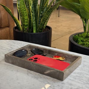 Op de afbeelding: Een rechthoekige grijze stenen tray met een zwarte zonnebril, een rode smartphone en een set sleutels. De tray staat op een wit marmeren oppervlak, met potplanten op de achtergrond.