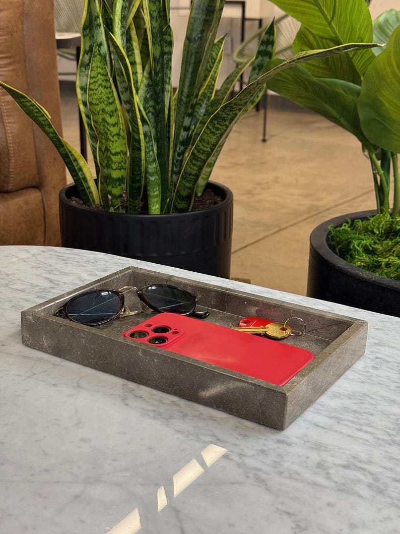 Op de afbeelding: Een rechthoekige grijze stenen tray met een zwarte zonnebril, een rode smartphone en een set sleutels. De tray staat op een wit marmeren oppervlak, met potplanten op de achtergrond.