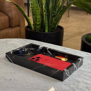 May include: A rectangular black marble tray with white veining holds a red smartphone, sunglasses, and a gold key. The tray sits on a white marble table. A potted plant is in the background.