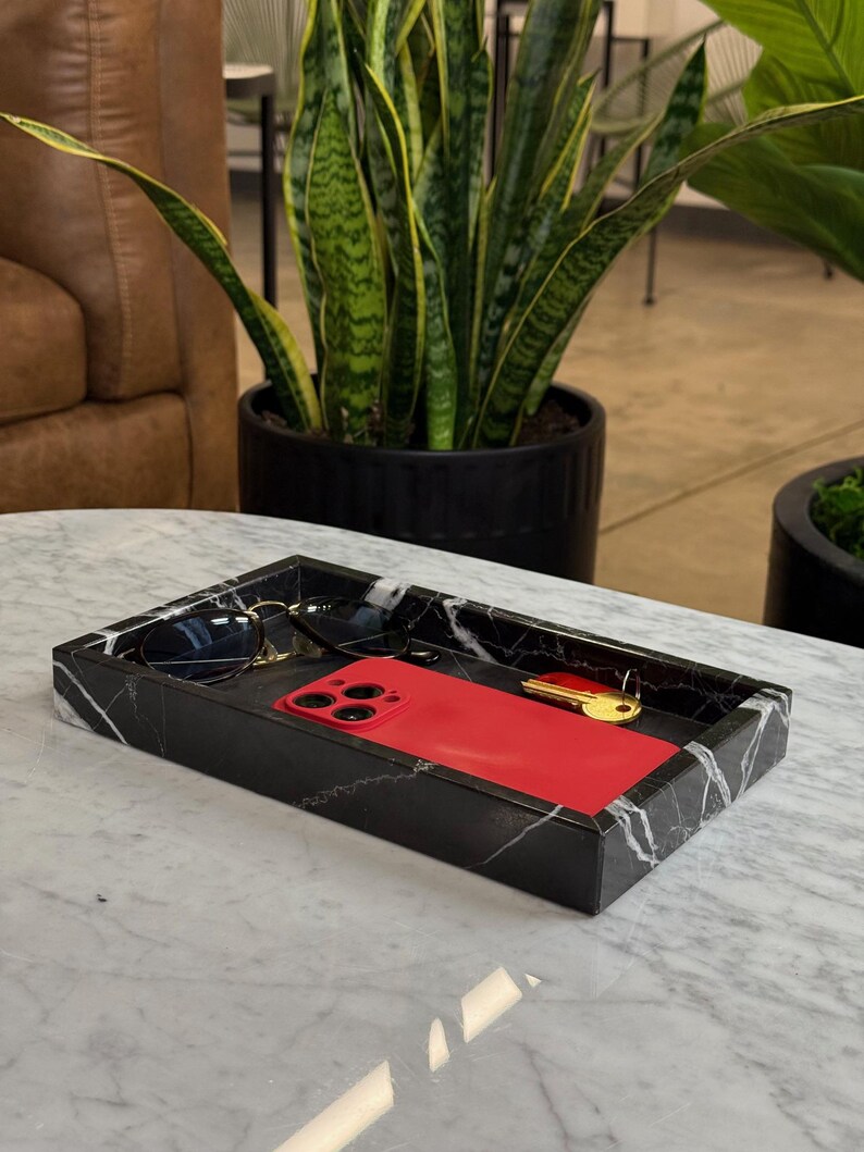 May include: A rectangular black marble tray with white veining holds a red smartphone, sunglasses, and a gold key. The tray sits on a white marble table. A potted plant is in the background.