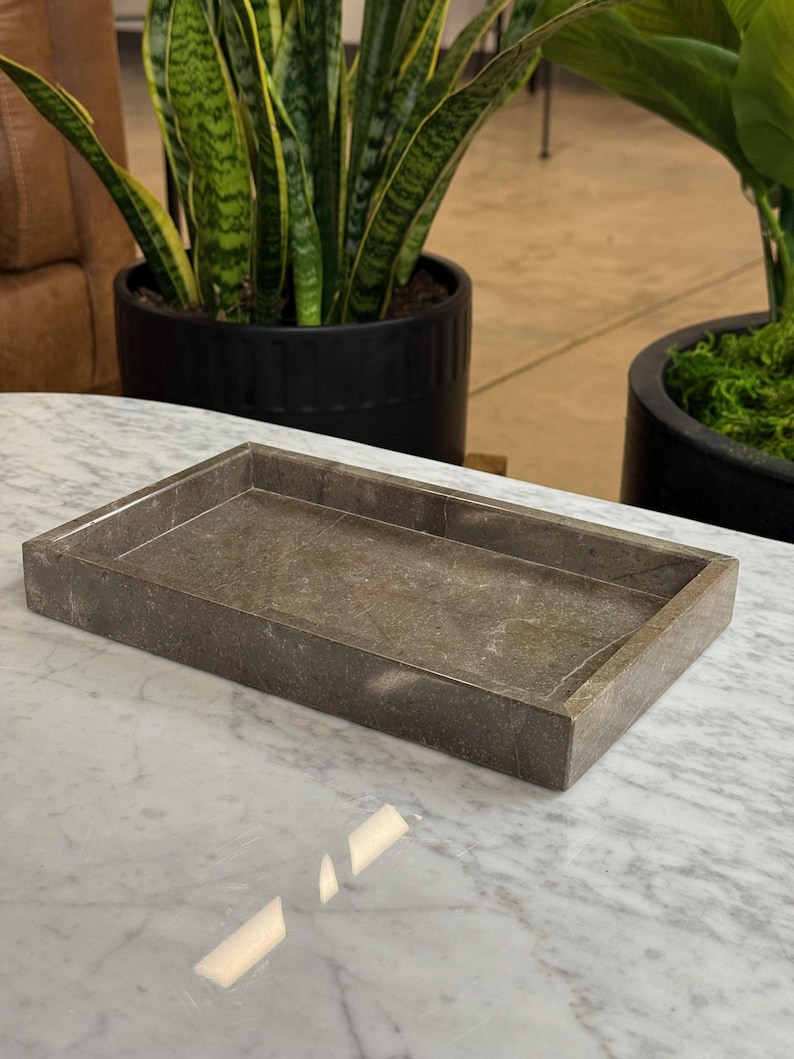 Op de afbeelding: Een rechthoekige, grijze marmeren dienblad met een opstaande rand. De lade staat op een wit marmeren oppervlak. De achtergrond bevat potplanten.