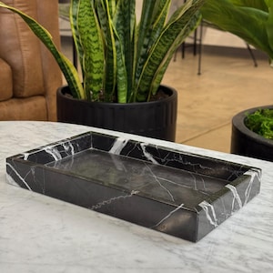May include: A rectangular black marble tray with white veining. The tray has a raised edge and a smooth interior surface. The tray is sitting on a white marble surface. The background includes potted plants.
